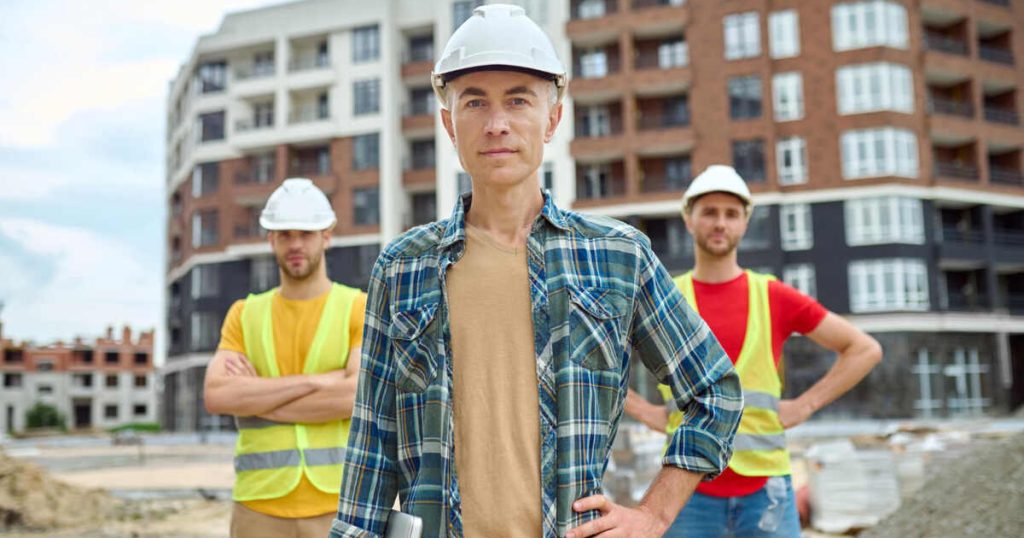 Three male construction workers standing in work clothes, one positioned in front, with a view of buildings under construction in the background.