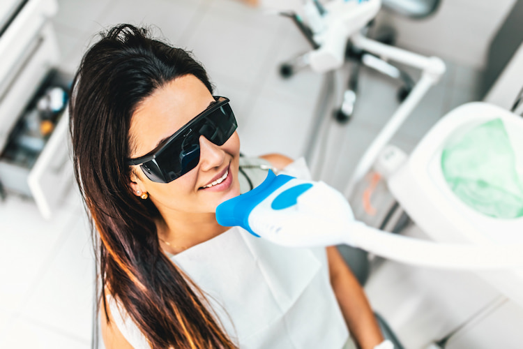 a woman is sitting in a dental office to get teeth whitening treatment while wearing protective glasses. 