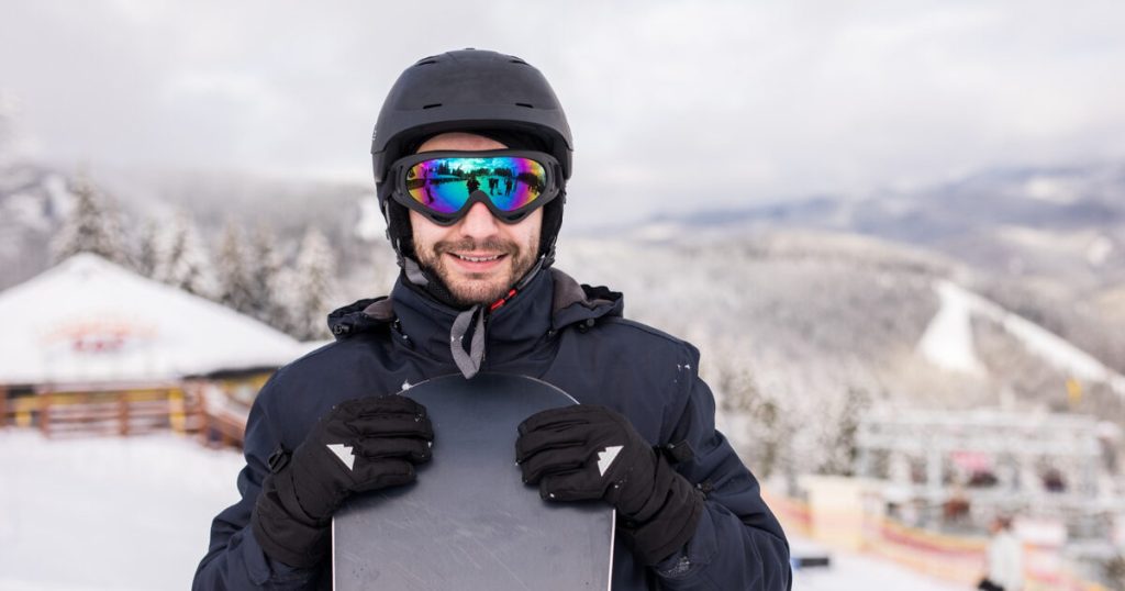 A smiling male snowboarder holding his snowboard in one hand, standing against a backdrop of snowy mountain peaks.
