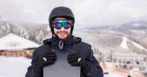 A smiling male snowboarder holding his snowboard in one hand, standing against a backdrop of snowy mountain peaks.