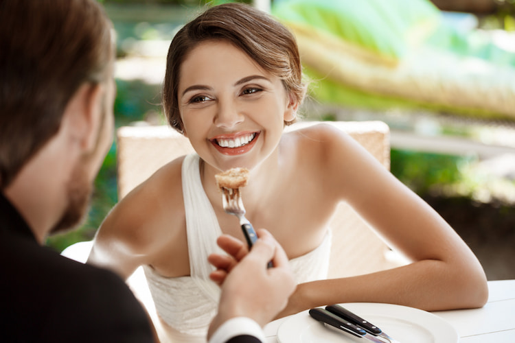 Smiling bride in white dress being fed a bite of food by a groom during a wedding party.