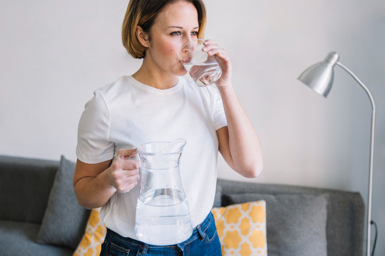 Young woman at home drinking water from a glass while holding a water pitcher.