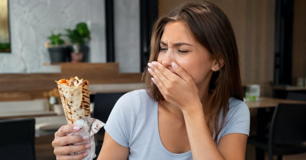 Young woman sitting in a restaurant holding shawarma and covering her mouth because of bad breath after eating shawarma