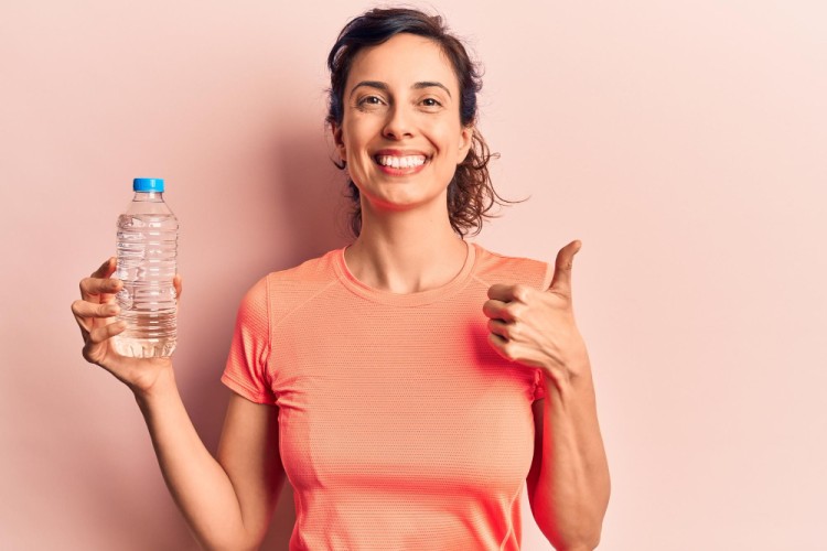 Young woman looking at the camera holding a water bottle in one hand and showing a thumbs-up with the other hand