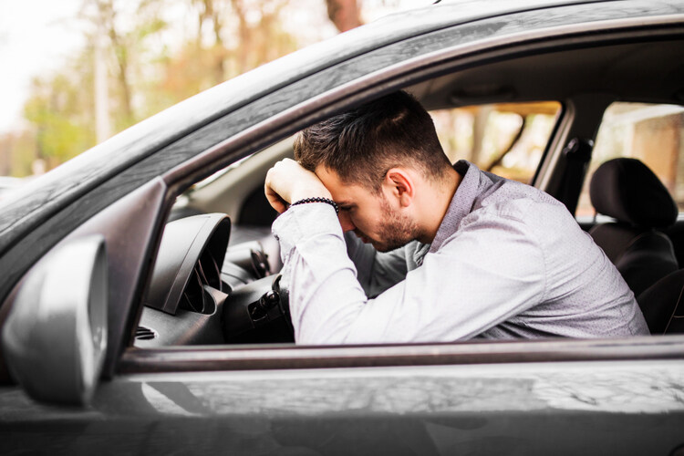 Frustrated man stuck in traffic with head resting on hand on steering wheel.