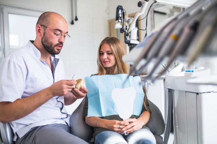 A male dentist explaining to a female patient whether a dental implant or bridge is the right option for her, while holding a dental model in his hand inside a modern dental clinic.
