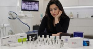 Young woman thoughtfully reviewing different dental implant brands displayed on a table in a dental clinic setting.