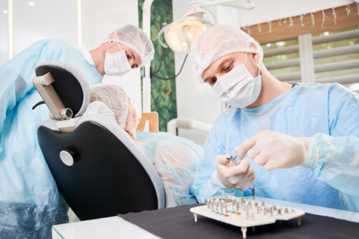 A male dentist and his assistant performing a dental implant procedure on a patient. The dentist is selecting the appropriate implant from a tray of dental implants on the table