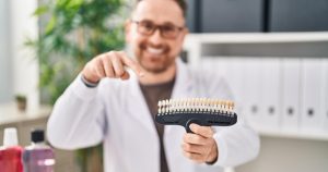 A smiling male dentist sitting behind his desk in a dental clinic, holding a row of porcelain veneers toward the camera.